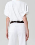 A person faces away, wearing the Citizens Of Humanity/AGOLDE gia bubble tee—a loose white jersey cotton short-sleeve top—tucked into high-waisted white jeans with a black belt. The background is plain and light-colored.