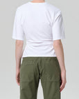 A person faces away from the camera, wearing the fabien tee by Citizens Of Humanity/AGOLDE—crafted from soft white jersey cotton—paired with green pants featuring large back pockets, set against a plain light gray backdrop.