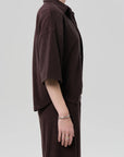 A person stands in profile against a plain background, wearing the Citizens Of Humanity/AGOLDE francis knit shirt in dark brown with short sleeves and a matching skirt. Their face is hidden, and they wear silver rings and a bracelet on their left hand.