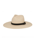 The Ninakuru Coda Hat, a beige wide-brimmed fedora made from toquilla straw, features a black band with a discreet bronze metal buckle around the crown, all set against a plain white background.