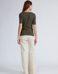 A woman with long brown hair, facing away, wears the knit contrast ringer tee by Rag & Bone in olive green and high-waisted cream wide-leg pants against a plain light gray background.