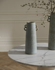 The Bowen Vase M by Accent Decor, a tall ceramic piece with handles and a soft blue glaze, sits on a round white marble table. Sparse leafy branches are arranged inside, with a cream paneled wall and wooden floor in the background.