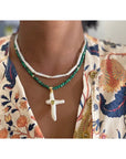 A person wearing a floral print shirt and two necklaces: one with malachite cubes and white beads, the other—the Carmelia Necklace by Faire—with green beads and a large peridot cross pearl pendant.