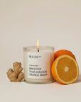 A lit Below 60 Candle by Faire with orange & ginger scent sits next to fresh ginger root and an orange, one half sliced open, on a neutral background.