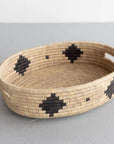 The Faire Palm Leaf Oval Basket is a shallow, handwoven tray with two handles and bold black geometric patterns, crafted by fair trade artisans. It rests on a light gray textured surface.
