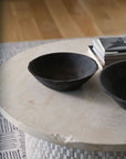 The Rossetti Bowl SM by Accent Decor in matte black sits on a round, light stone table beside books, with another handcrafted terracotta bowl and a patterned rug visible in the background.
