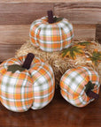 Three Faire Pumpkin decorations in orange, green, and white plaid rest on a wooden surface—two on the table and one on a hay bale with autumn leaves—adding cozy farmhouse fall charm against a wood panel backdrop.