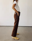 A person stands sideways by a plain wall, wearing a white t-shirt and Hey Gang's The Work Pant in brown cotton canvas with cropped legs, paired with light tan boots. Their left hand is in their pocket and their face isn't visible.