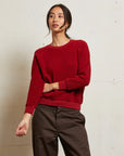 A woman with long dark hair pulled back stands against a light wall, wearing Perfectwhitetee's Ziggy Reverse LS Sweatshirt in a shrunken fit and brown pants, resting one hand on her opposite arm and gazing slightly to the side.