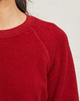 Close-up of a person wearing the Perfectwhitetee Ziggy Reverse LS Sweatshirt in textured red, showcasing its shrunken fit and crew neckline. The neutral background highlights the sweater's details around the shoulder and upper chest.