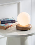 The Faire Valencia Portable Light with a wooden base rests on a small white table beside two stacked books, including one by Sidney Sheldon, creating a bright and minimalistic scene.