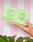 A manicured hand displays a green Faire Cucumber/Tea Eye Mask package against a pink and white striped backdrop, emphasizing its natural ingredients. Green leaves are partly visible at the bottom.