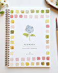The Faire Watercolor Workbook, spiral-bound and beginner-friendly, features a blue hydrangea cover with colorful borders and sits near a paintbrush, palette, and white flowers—perfect for aspiring artists.