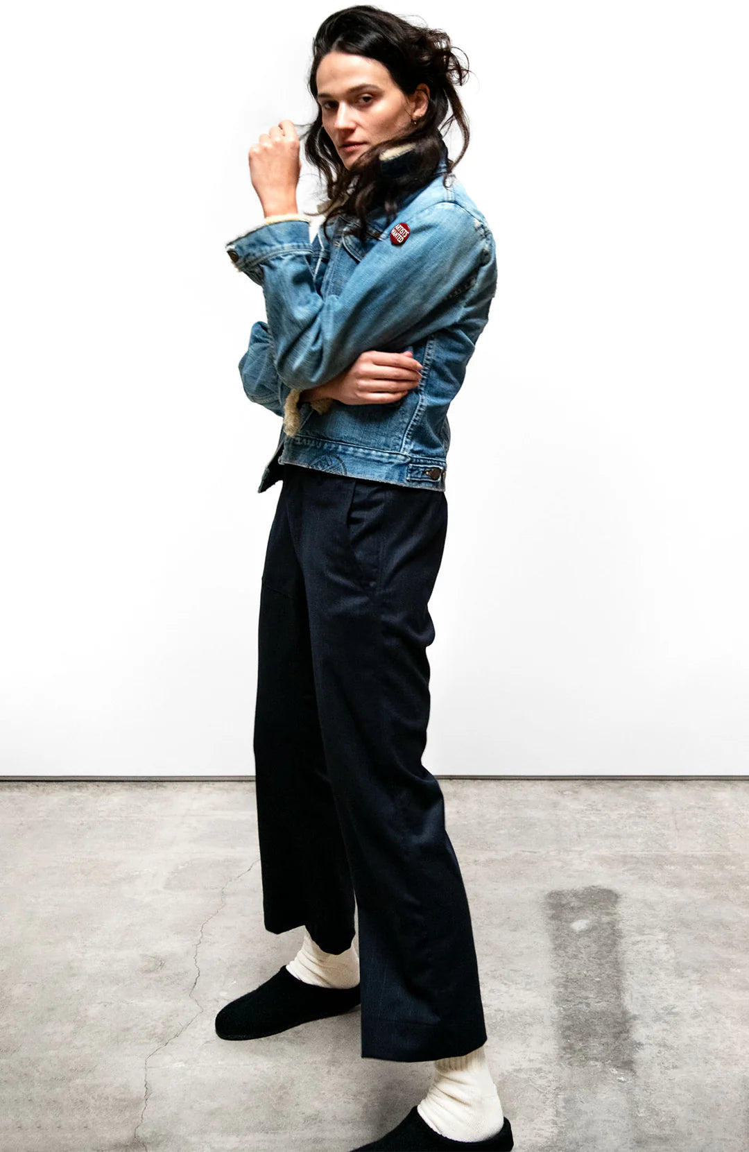 A person with long dark hair stands indoors on a concrete floor, wearing a denim jacket, Free City Favolamove Cashmere French Sailor Pant, white socks, and black slip-on shoes, posing with one arm bent and hand near their face.