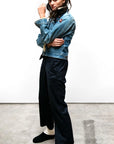 A person with long dark hair stands indoors on a concrete floor, wearing a denim jacket, Free City Favolamove Cashmere French Sailor Pant, white socks, and black slip-on shoes, posing with one arm bent and hand near their face.