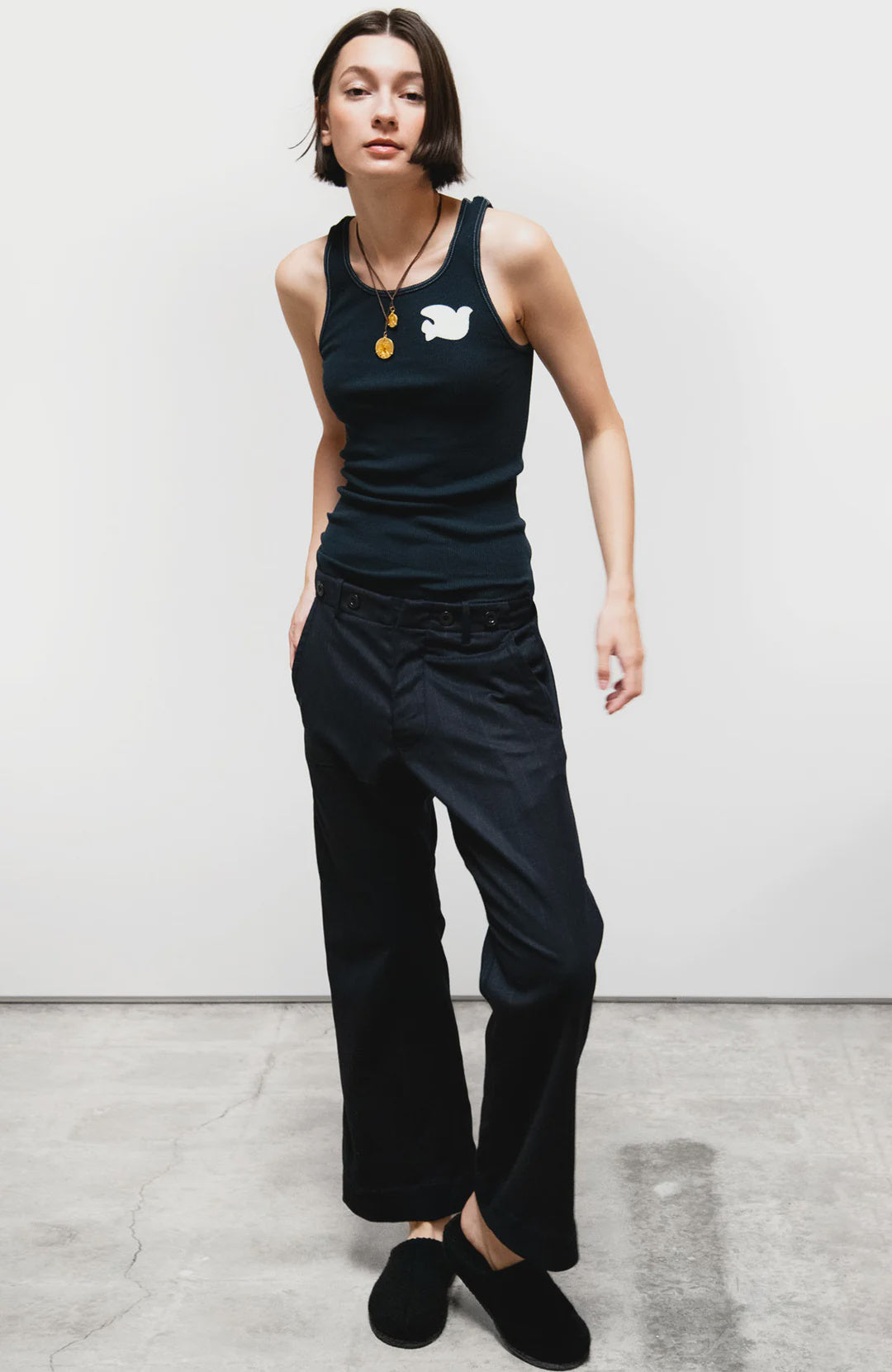 A person stands against a plain white wall wearing a sleeveless black tank top with a white graphic, black shoes, a round pendant necklace, and Free City Favolamove Cashmere French Sailor Pant. Their hands are in their pockets.