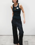 A person stands against a plain white wall wearing a sleeveless black tank top with a white graphic, black shoes, a round pendant necklace, and Free City Favolamove Cashmere French Sailor Pant. Their hands are in their pockets.