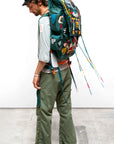 A man in a black cap, light shirt, and Free City CanyonKid Tanks Flap/Snap Homestud pant stands sideways, wearing a large colorful backpack with long beaded tassels against a plain background. MADE IN USA.