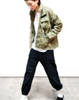 A person in an olive green jacket, white t-shirt, and Free City (sparrow, LLC) Cashmere/Wool Flap/Snap Pant walks indoors on a concrete floor against a white background.
