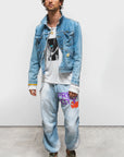 A man stands against a plain background wearing the Free City (sparrow, LLC) CanyonKid Home/Stud Flap/Snap, a graphic tee, light blue jeans with “FREE CITY” in purple and orange print, green sneakers, black cap, necklace, and bracelet.