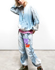 A person with blonde hair wears a light blue, oversized Free City CanyonKid Home/Stud Flap/Snap sweatsuit with FREECITY print, Unisex INDIGO pant featuring “FREEDY” and a red graphic, pink shirt underneath, green sneakers, and layered necklaces.