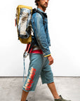 A man wearing a denim jacket, Free City (sparrow, LLC) Sparrow Homestud Yumm Cutoffs with red and white graphics, green sneakers, a black cap, and a large handmade backpack walks across a concrete floor against a plain white background.