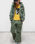 Dressed in a Free City (sparrow, LLC) CanyonKid Tanks Flap/Snap Homestud, black cap, green jacket, yellow graphic tee, Unisex Karate Pants, and blue sneakers, the individual stands against white with a Vintage Army Bag and hands in pockets.