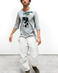 A man in a black cap, light gray graphic long-sleeve shirt, and Free City Out of Sight Poplin Jump Pant in custom laundry whites with green sneakers stands on a concrete floor against a plain white wall.
