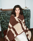 A woman with wavy dark hair sits on a chair, wrapped in the Autumn Sonata Karin Bath Sheet. She wears a white skirt and looks at the camera, with a dark green stone fireplace and white bowl visible in the background.