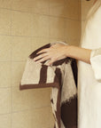 Wearing a cream robe, a person holds the Autumn Sonata Karin Hand Towel—featuring brown and beige stripes—in front of a beige tiled wall. Only part of their face and body is visible.