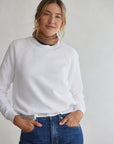 A woman with blonde hair in a loose ponytail wears the Perfectwhitetee Ziggy Reverse LS Sweatshirt and blue jeans, paired with a brown and gold choker necklace, standing against a light gray background with her hands in her pockets.