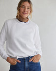 A woman with blonde hair in a loose ponytail wears the Perfectwhitetee Ziggy Reverse LS Sweatshirt and blue jeans, paired with a brown and gold choker necklace, standing against a light gray background with her hands in her pockets.