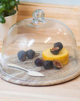 The Be Home Waterfall Marble Cloche features a round pink and teal marble cheese board with a glass dome, showcasing yellow cheese and figs with a spreader for stunning artisanal decor on your wooden countertop.