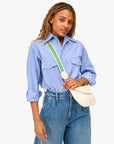 A person with long hair wears rolled-up sleeves on a blue shirt and jeans, accessorizing with Clare Vivier's white quilted crossbody bag featuring the Crossbody Strap S25 in green with brass snap hooks. The background is plain white.