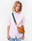 A person wearing a light pink button-up and blue jeans poses with the Clare Vivier Adjustable Crossbody Strap, a woven brown bag with a thick green cotton webbing strap, against a plain white background.