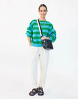 A woman smiles against a white background, wearing a green and blue striped sweater, white cuffed pants, black sandals, and a black Clare Vivier Crossbody Strap S24 bag with a nylon webbing strap. She accessorizes with simple jewelry.