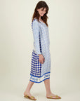 A woman with long brown hair, wearing the Me369 Violet V-Neck Tee Dress in blue and white with a geometric print and gold sandals, stands sideways and smiles. The dress hem features the words "Enjoy" and "Just.