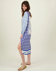 A smiling woman stands sideways in a Me369 Violet V-Neck Tee Dress with “Just Enjoy” near the hem. She pairs the blue and white geometric print dress with gold sandals, her long brown hair flowing against a plain, light background.