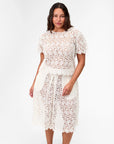 A woman stands against a plain white background wearing the Clare Vivier Lou Lou Top, a short-sleeved, white cotton lace top with floral pattern and see-thru design.