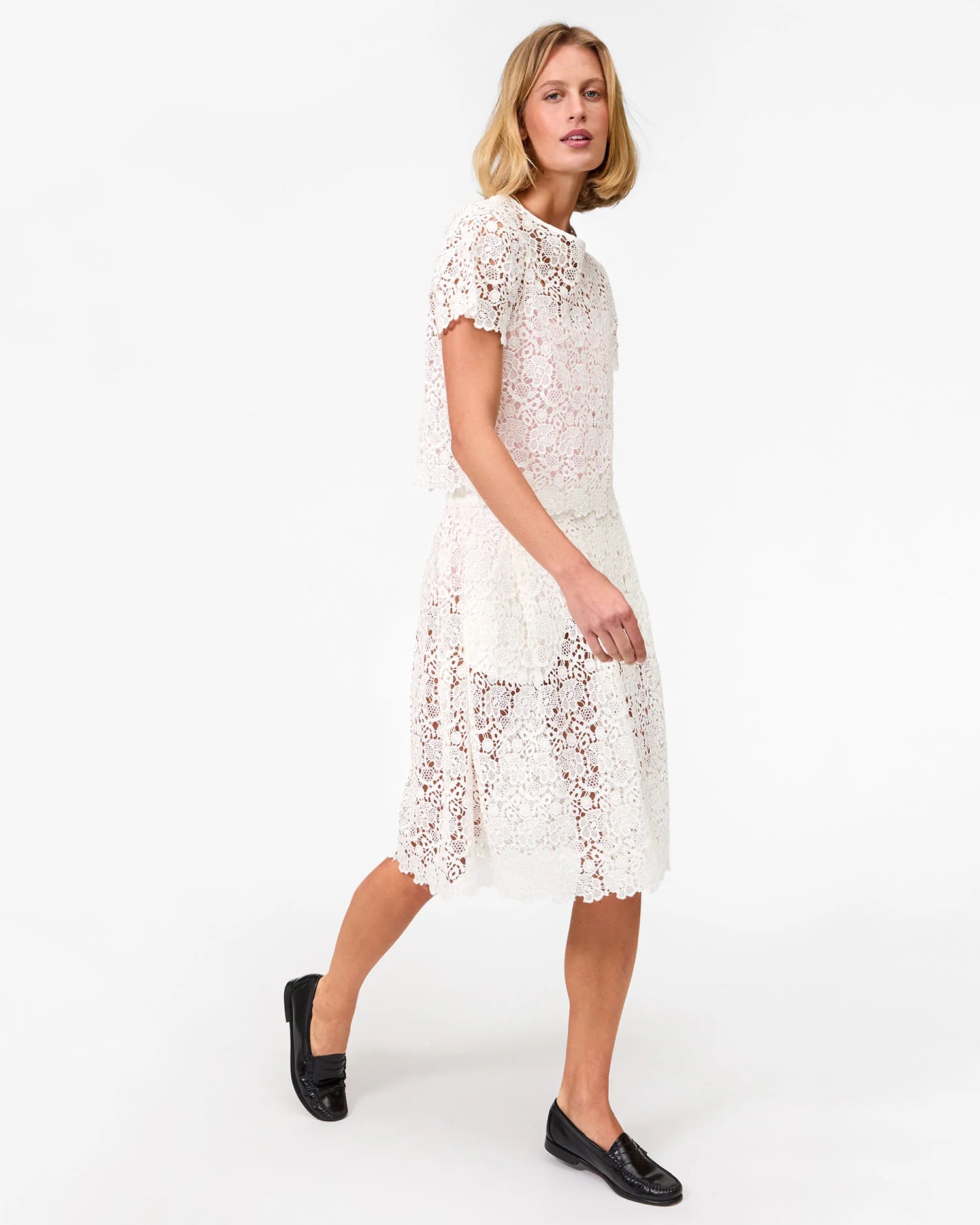 A woman with blonde hair wears the Clare Vivier Anais Court Skirt and black loafers as she walks against a plain white background.
