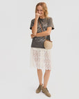 A smiling woman stands against a white background wearing the Clare Vivier Anais Court Skirt, a gray graphic t-shirt, tan ankle boots, and a round woven crossbody bag, touching her face with one hand.