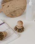 Two Be Home Saucepan Brushes with vegetable fiber bristles rest on a white countertop beside a folded towel, round cutting board, and soap dispenser near the kitchen sink.