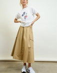 A woman stands against a plain background wearing a white Mickey Mouse t-shirt, white sneakers, and the Aquarius Cocktail Le Cargo Skirt—a tan Japanese cotton poplin skirt with oversized pockets. She poses relaxedly with one hand on her hip.