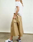 Someone stands sideways on a concrete floor against a plain light background, wearing a white t-shirt, white Nike sneakers, and the Le Cargo Skirt by Aquarius Cocktail—tan with oversized pockets in Japanese cotton poplin.
