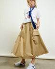 A person in a white Mickey Mouse t-shirt, blue tie, Le Cargo Skirt by Aquarius Cocktail with oversized pockets, and white Nike sneakers stands against a plain backdrop, looking down with hair partially covering their face.