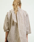 A person with long, wavy blonde hair styled as a face framer wears the loose-fitting Dear Polly Bow Blouse by Aquarius Cocktail, featuring a light base with a small brown spotted pattern. The photo is taken from the back against a plain background.