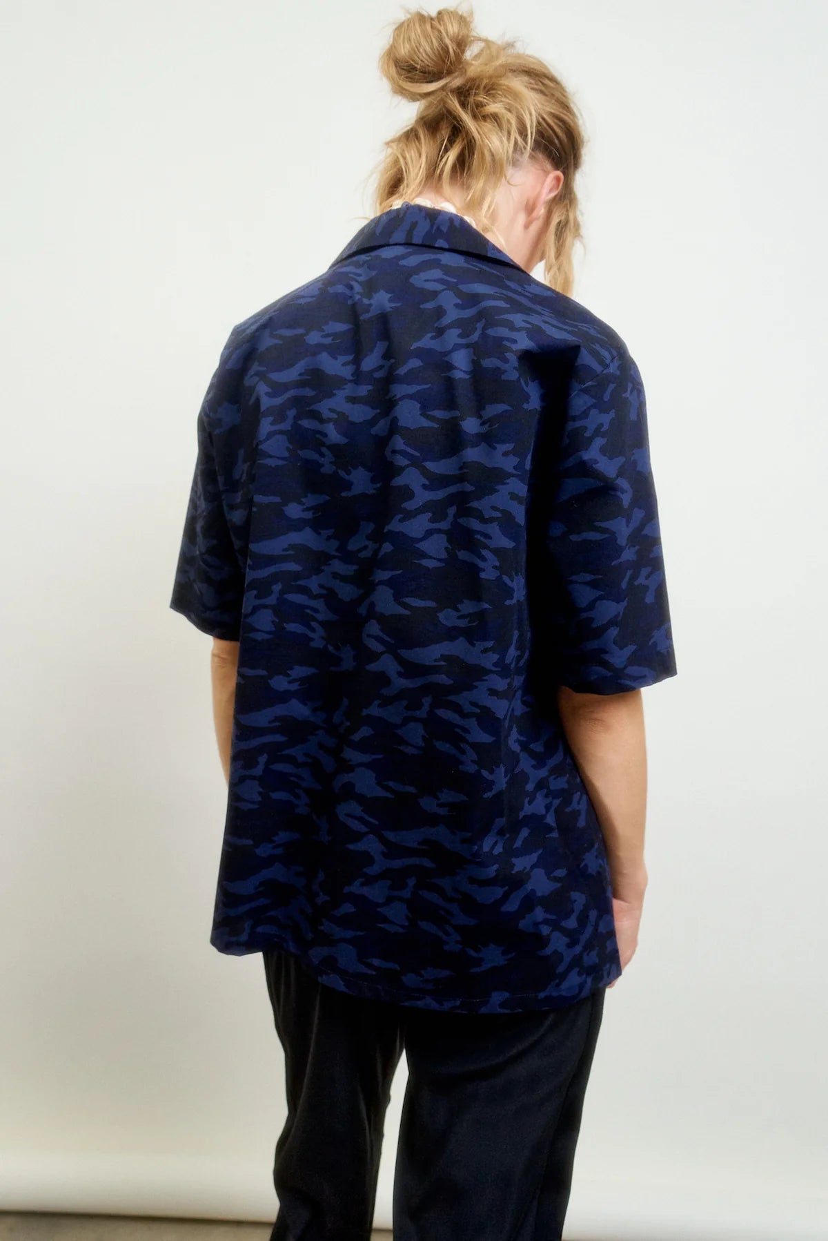 A person with light brown hair in a messy bun wears the Aquarius Cocktail CAMO LE Camp Shirt, featuring navy blue and black Japanese cotton, and stands facing away from the camera against a plain light background.
