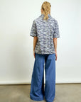 A person with long blonde hair in a ponytail stands facing away, wearing a grey camo short-sleeve shirt and the Aquarius Cocktail Connor Wide Leg Pant with an elastic waist, small batch made, against a white backdrop.