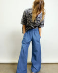 A person with long, light brown hair looks down, wearing a gray patterned shirt and Aquarius Cocktail’s Connor Wide Leg Pant in blue with an elastic waist, paired with beige shoes with white polka dots, standing against a plain light background.