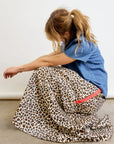 A person with light brown hair in a ponytail sits on the floor against a plain light background, wearing a blue short-sleeve shirt and the Aquarius Cocktail Le Cargo Skirt—a long leopard-print cargo skirt with oversized pockets and a pink stripe.